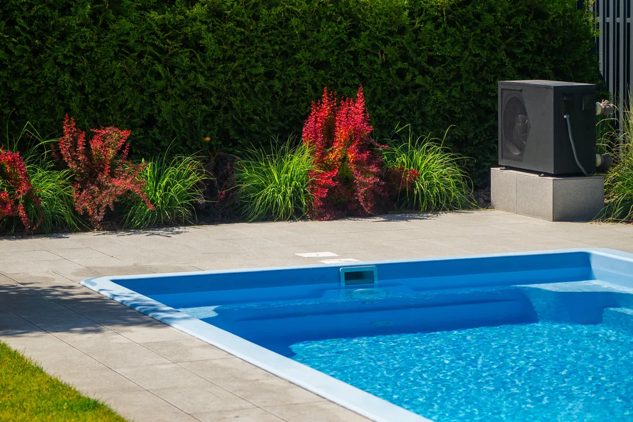 A paved stone patio wrapping the corner of an in-ground pool, with planting beds along a clipped hedge and a heat pump set on a block pad