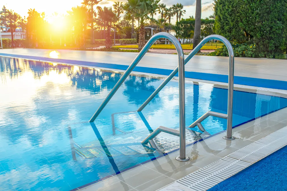 A long outdoor pool at low sun, with stainless handrails and entry steps at the near corner and light reflecting across the water
