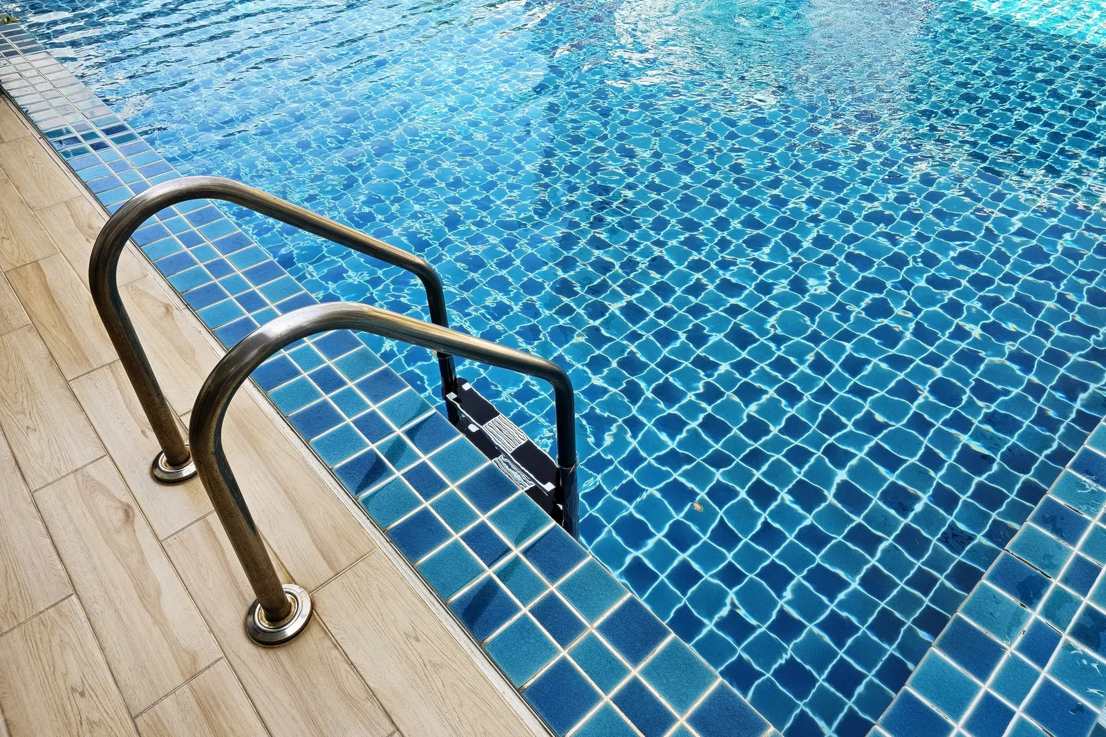 Stainless grab rails at the tiled edge of a swimming pool, with sunlit water over blue mosaic tile and a wood-look deck along the near side